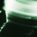 Imagem abstrata com tons esverdeados mostrando o perfil de um rosto parcialmente iluminado à esquerda, com rastros de luz dinâmica se espalhando pelo fundo escuro, criando um efeito etéreo e futurista.