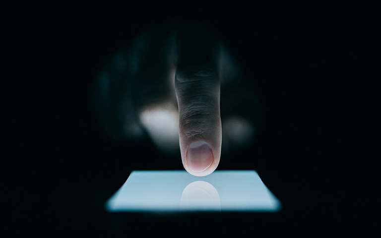 Dedo tocando a tela iluminada de um smartphone em ambiente escuro, destacando o contraste entre a luz do aparelho e o fundo preto