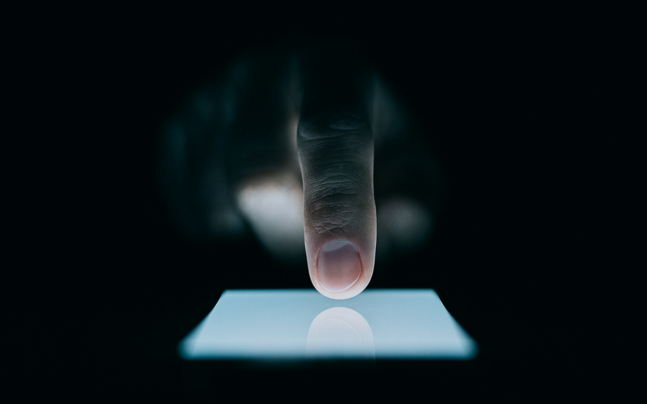 Dedo tocando a tela iluminada de um smartphone em ambiente escuro, destacando o contraste entre a luz do aparelho e o fundo preto