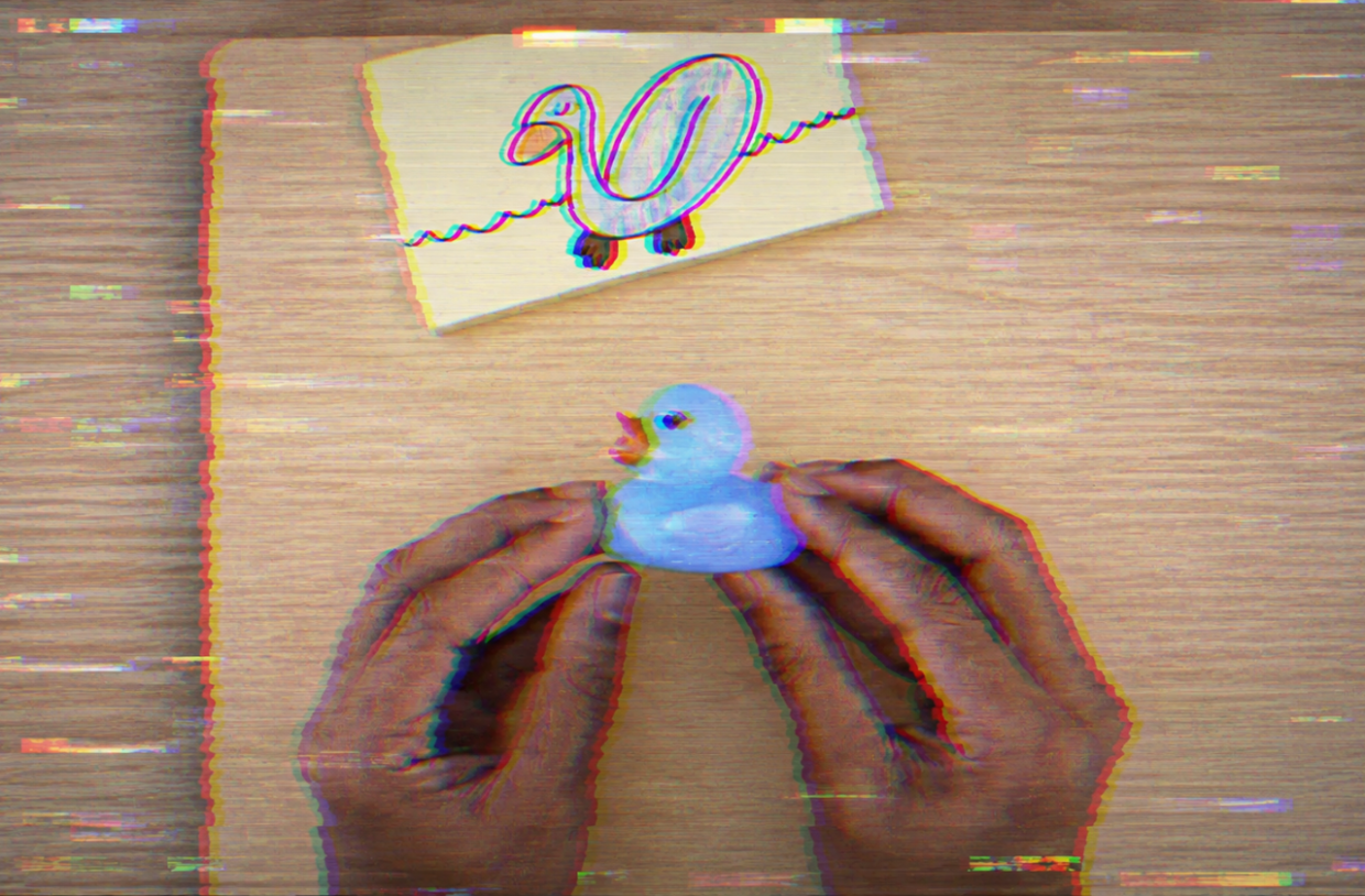 Mãos seguram um pato de borracha azul sobre uma mesa de madeira clara, abaixo de um desenho simples de um pato em papel. A imagem em formato 16:9 apresenta forte efeito de falha digital, com serrilhado, distorções RGB, scanlines e glitches que transmitem a ideia de corrupção visual.